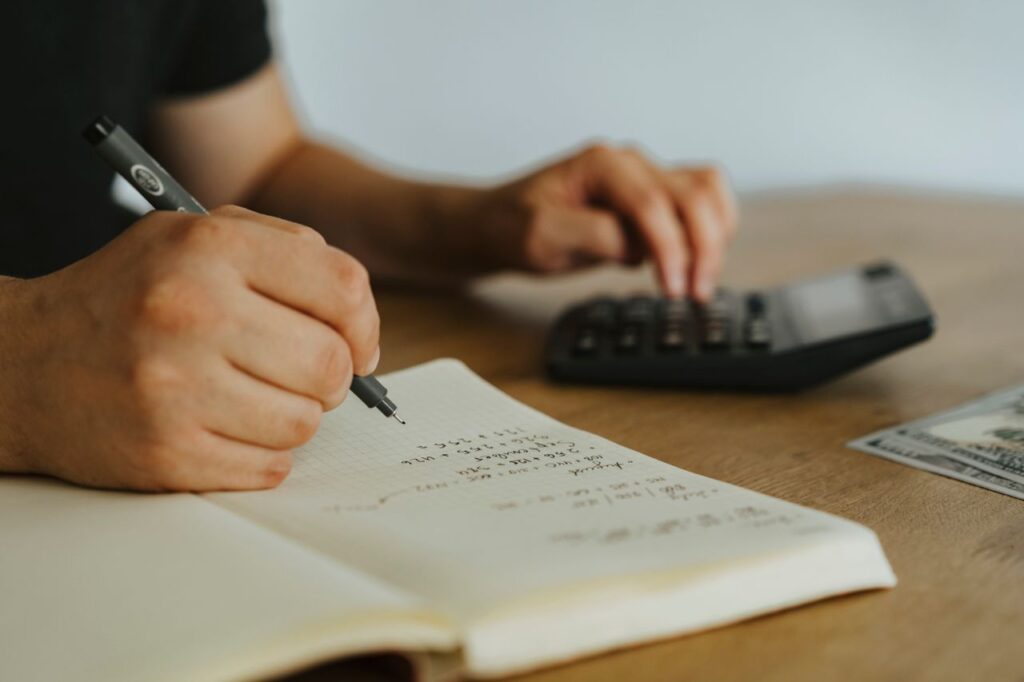 Bookkeeping Services Across the United States Close-up of hands writing calculations in a notebook with a calculator, focused on budgeting or financial work.